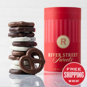 A red cylindrical canister with the logo 'RIVER STREET Sweets' on it, alongside a stack of assorted chocolate-covered pretzels.