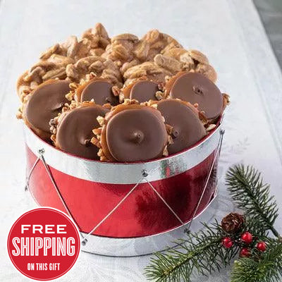A red cylindrical drum-shaped box containing an assortment of pralines and bear claws, with holiday decorations in the background and a 'Free Shipping' badge displayed.