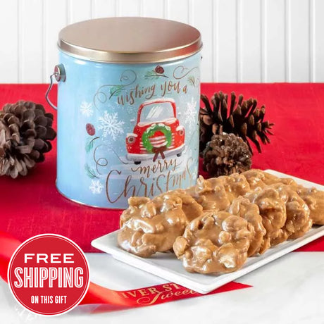 A festive tin of Savannah's World Famous Pralines with a snowy background, decorated with Christmas elements, accompanied by a plate of praline sweets. The tin has text and a graphic of a red truck with 'Merry Christmas'.