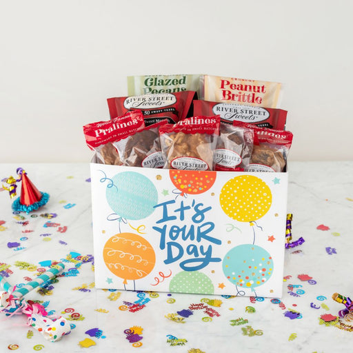 Gift box with snacks and a card saying 'It's Your Day' on a celebratory background.
